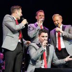 Four men in grey blazers and red ties singing into hand mics, one kneeling before the other three