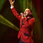 White man in red glittery jacket sings, one hand thrown in the air.