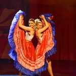 Two women in cropped tops holding their long red skirts with blue fringing up above their heads as they stand back to back.