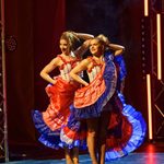 Two women in long red skirts with blue fringing up flick their skirts around as they hold one hand behind their head, standing in line.