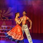 White woman in long red skirt with blue fringing turns to bare chested man in white trousers and red suspenders as they dance.