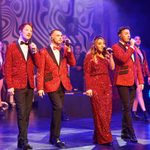 3 men and 1 woman in red glittery out fits sing together.