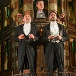 2 men standing on balcony set holding glasses of wine, young man standing inbetween behind holding bottle of wine.