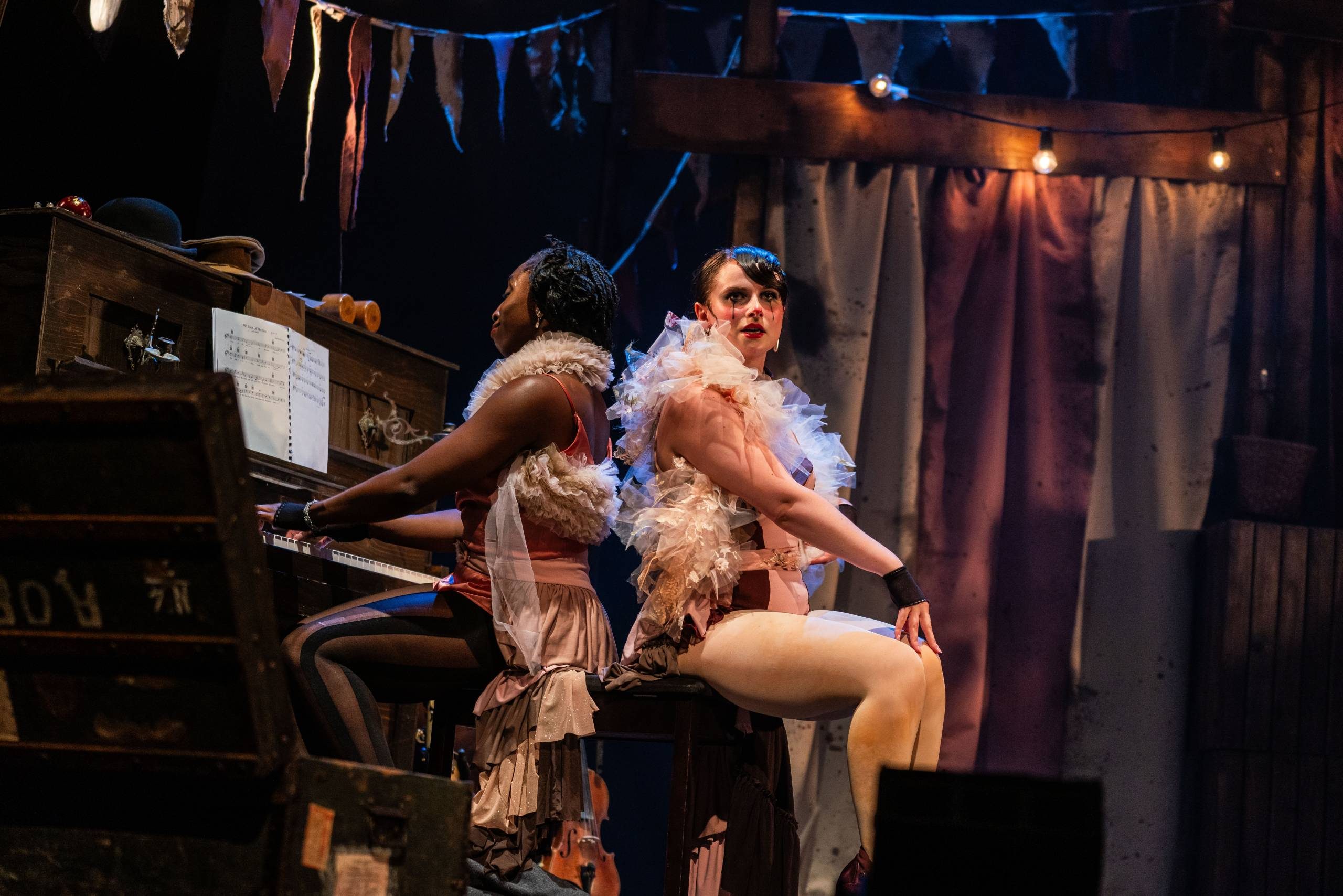 Two females sit back to back, One plays the piano and the other sits towards the audience.