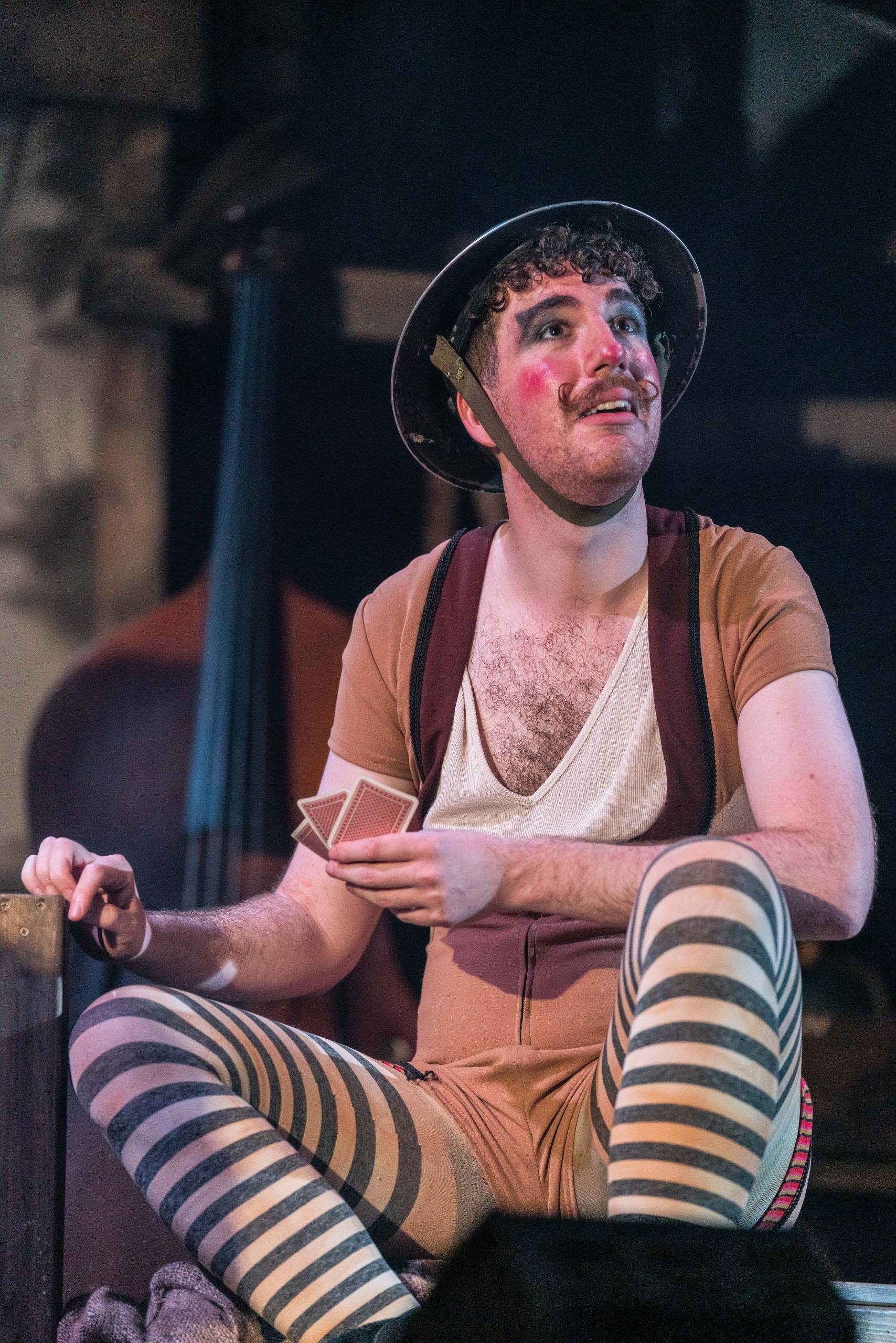 a man wearing stripy tights, brown top, face makeup and soldiers hat looks up.