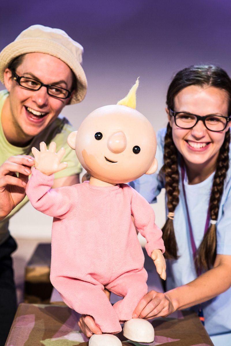 A baby puppet is waving, the puppet is operated by a male and female character.