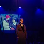 A young white female performing under a spotlight whilst images from our Bugsy Malone photoshoot rotate on a screen behind her.