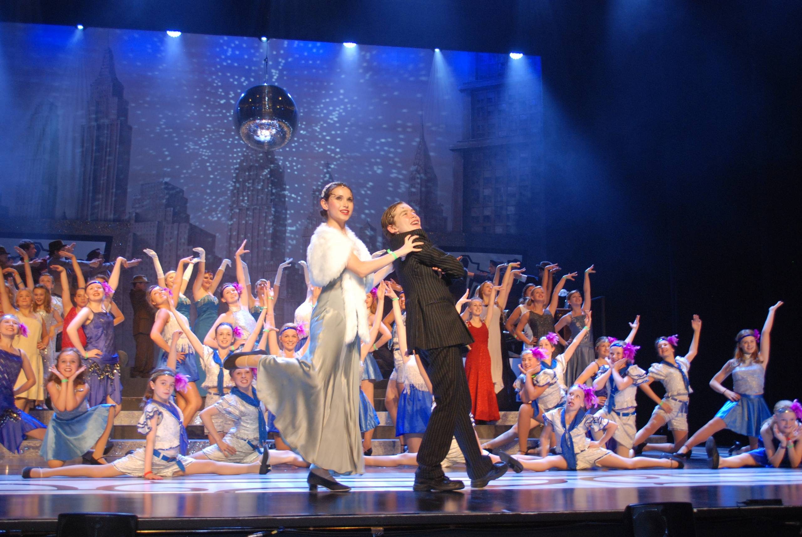 The cast of Bugsy Malone pose with arms in the air. There is a city backdrop and a disco ball hanging.