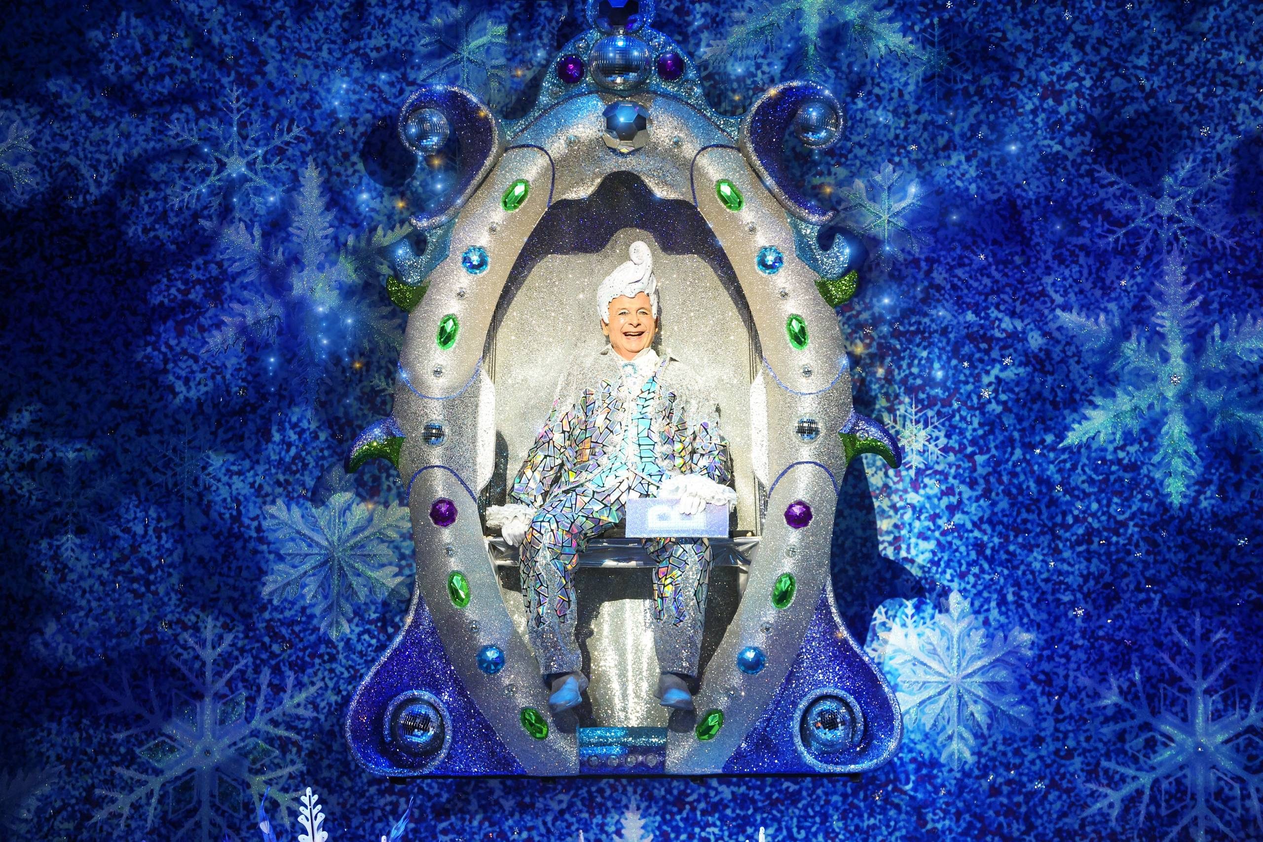 Christopher Biggins sits in a sparkly mirror set with a blue snowflake background behind him