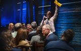 A male holds a yellow envelope in the air while speaking into a phone. Ten other visible cast members are sat on chairs smiling and laughing.