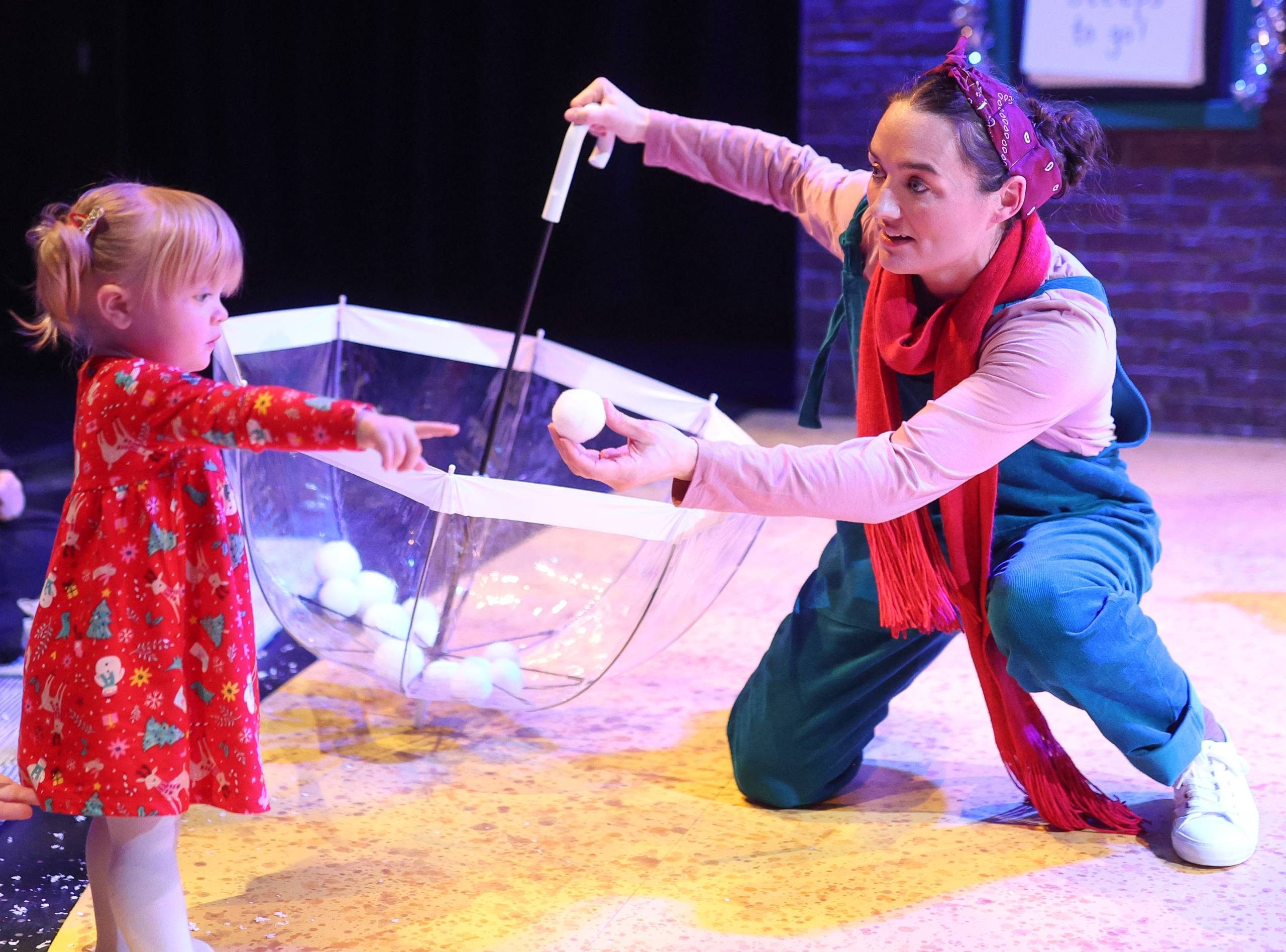 Female character holding an umbrellas passes a snowball to a young child.