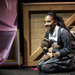 A school girl crouches in front of a wooden box clutching a teddy bear.