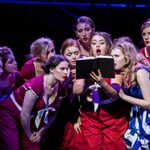 Young female actors singing on stage whilst looking at a book from our 2017 Summer Youth Project Guys and Dolls.