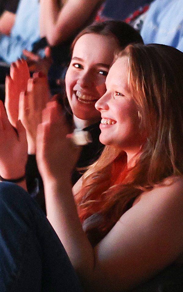 Close up image of a young white girl sat in the auditorium smiling snd clapping her hands.