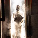 A smartly dressed male character emerges from a doorway brightly lit with white light.