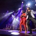 Black woman dressed all in red and black man in silver jacket singing back to back in foreground left of stage, a mirroring in the background to the right of the stage.