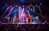 Five singers, two black men in silver jackets with 3 black women all in red with short capes between them, a band dressed in white behind, the crowd in shadow looking up at them