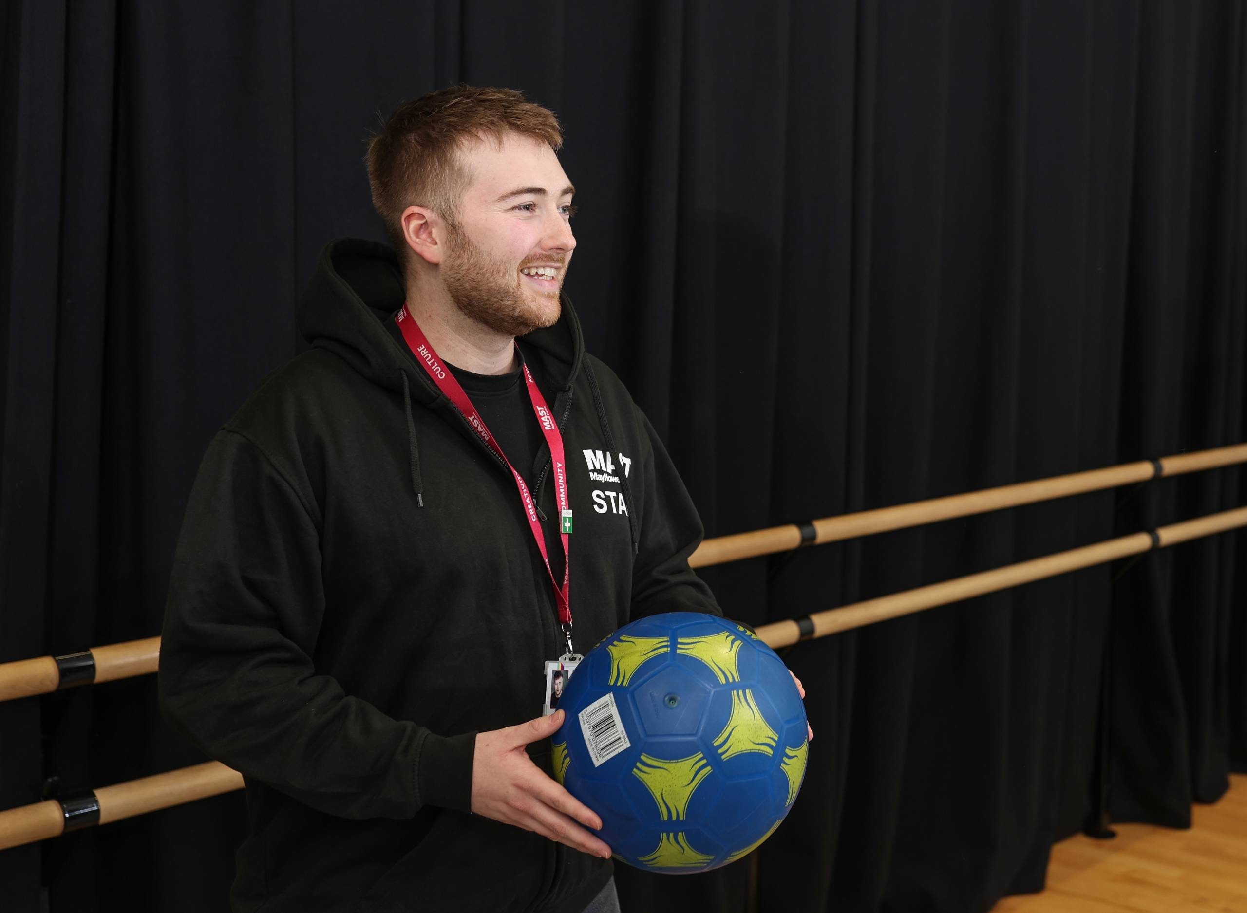 A man in a black MAST staff hoodie is holding a blue and yellow football and smiling to someone out of frame.