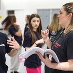 A female member of our engage team holds a script and directs a young participant. 5 other participants can be seen in the background.