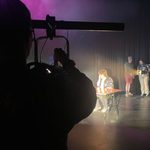 Close up image of a young person in silhouette shining a spotlight at performers on stage.