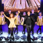 White woman in gold dress and white man in black suit raise hands to bow as behind them a line in the same outfits stands hand in hand.