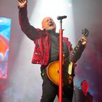 Steve Steinman in red leopard print jacket, holding wood-effect electric guitar, passionately throwing a hand up as he sings.