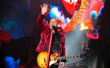 Steve Steinman in red leopard print jacket, holding wood-effect electric guitar, one hand raised as he passionately sings.