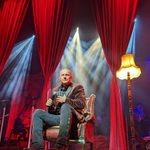 Steve Steinman in leather arm chair, lit by standing lampshade, long red curtains drawn behind him