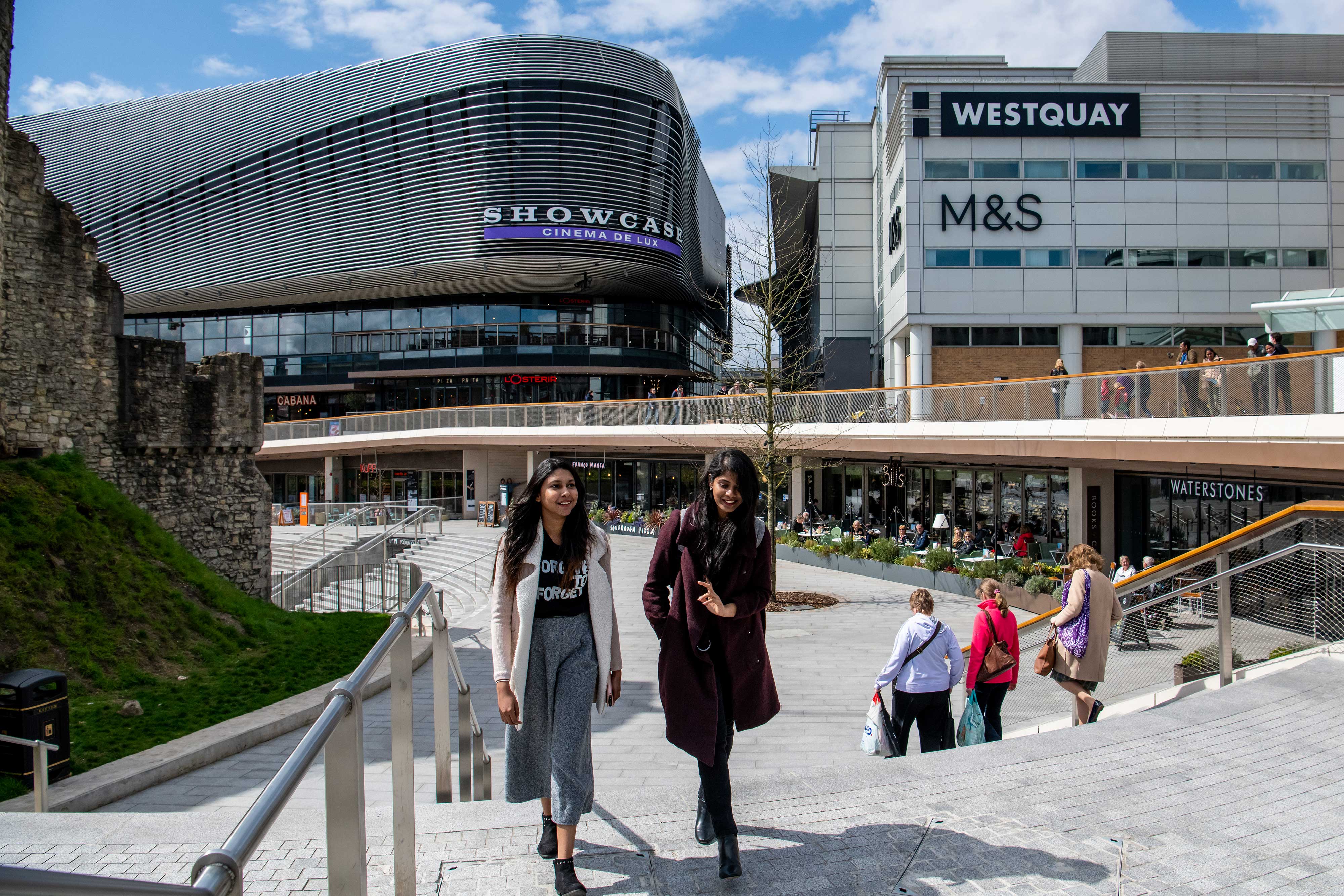 Two students talking whilst crossing a large paved plaza surrounded by shops, on a sunny day.