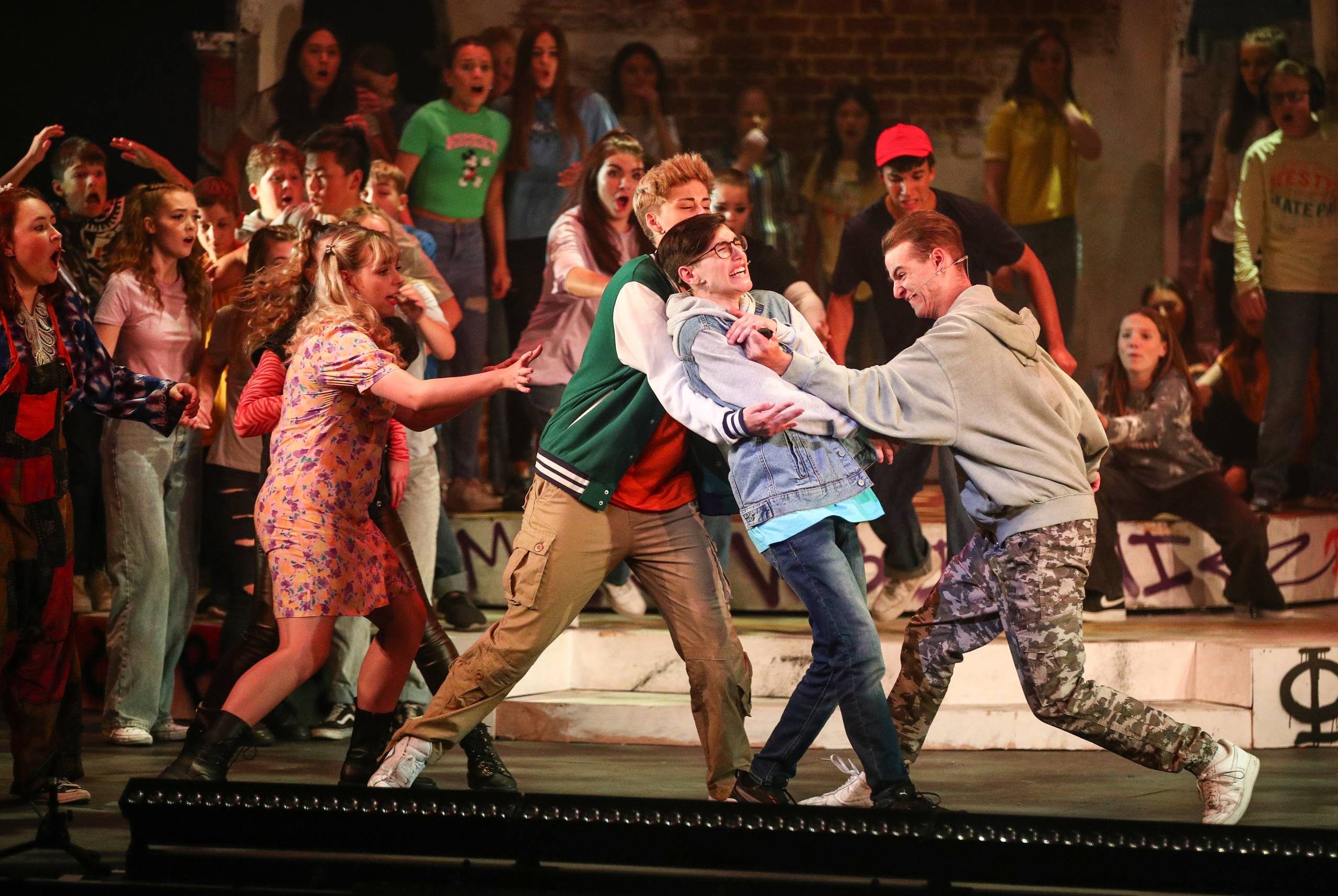 The main cast member is being attacked by the villain of the show whilst the ensemble stand behind and watch in horror.