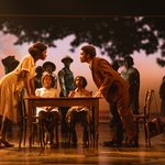 A black man and woman lean over a table above two young black girls, ensemble watching on from shadowed trees to back of stage.