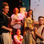 A group of young white male and female performers. The male is holding a shoe in the air and confetti is falling around him.