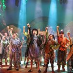 A diverse group of young people performing on stage with their fist raised in the air.