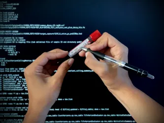 Close-up of hands labelling a patient blood sample for molecular analysis. In the background a computer screen shows programme code to process sequencing data. 