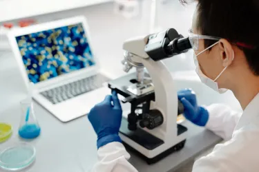 Student in a lab coat is looking into a microscope and showing results on the laptop infront of her