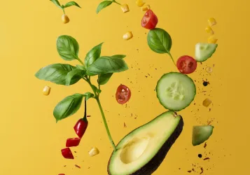 Half an avocado, halved cherry tomatoes, a red sliced chili pepper, basil leaves, cucumber slices and fine herbs fly into the air against a yellow background