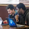 2 students at a cafe table chatting with laptops.