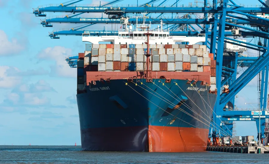 A heavily laden cargo ship sits under cranes by the dockside